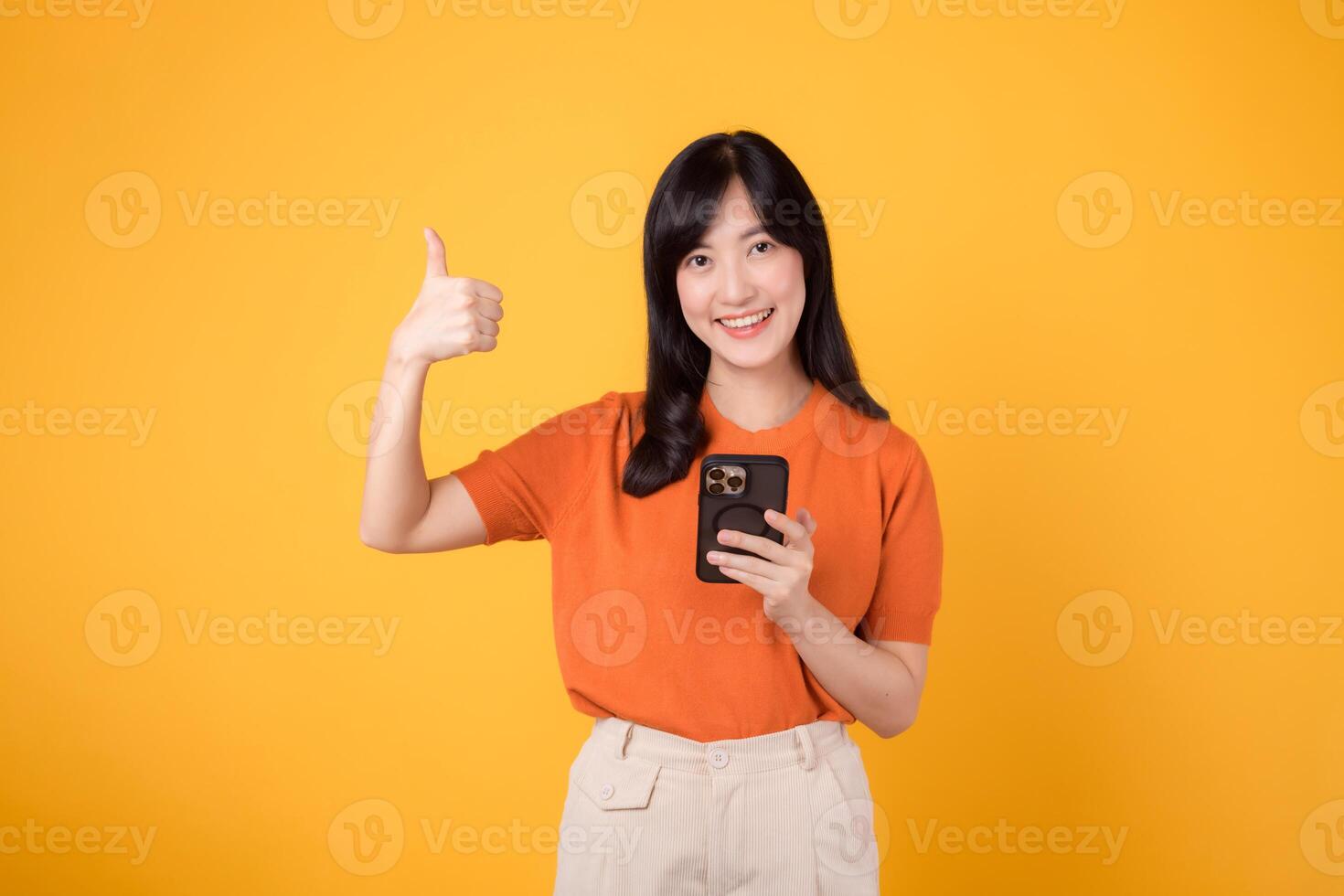 sorridente asiatico donna nel sua anni '30, indossare arancia camicia, utilizzando smartphone con pollice su mano cartello su vivace giallo sfondo. nuovo mobile App concetto. foto