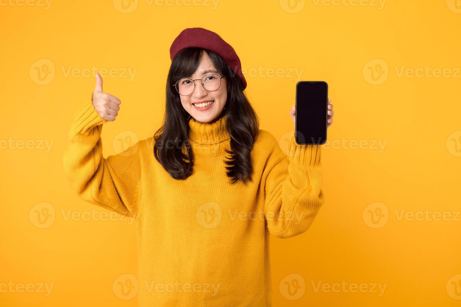 eccitato giovane asiatico donna 30s indossare giallo maglione, rosso berretto e occhiali mostrando mobile Telefono schermo, pollici su, piace Buona, consigliare smartphone applicazione, in piedi al di sopra di giallo sfondo foto