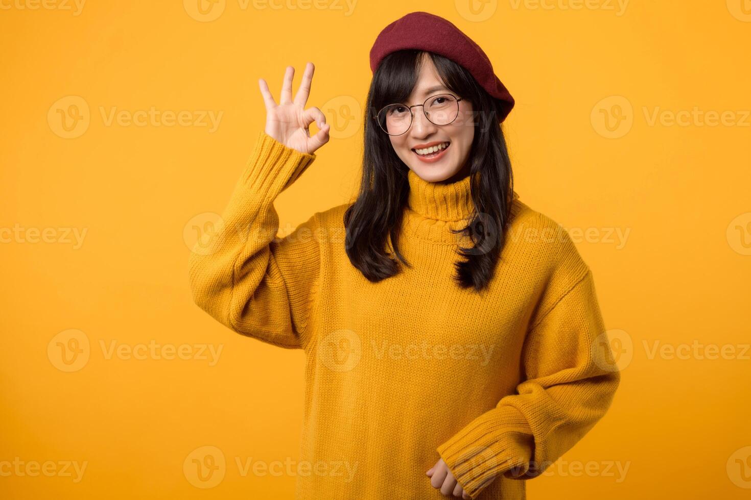 fiducioso donna nel un' giallo maglione, rosso berretto, e occhiali, fabbricazione il va bene gesto contro un' vivace giallo sfondo. foto