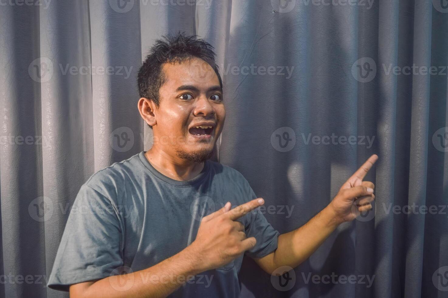 Wow e sorpreso viso di asiatico uomo nel grigio maglietta con mano punti su vuoto spazio. foto