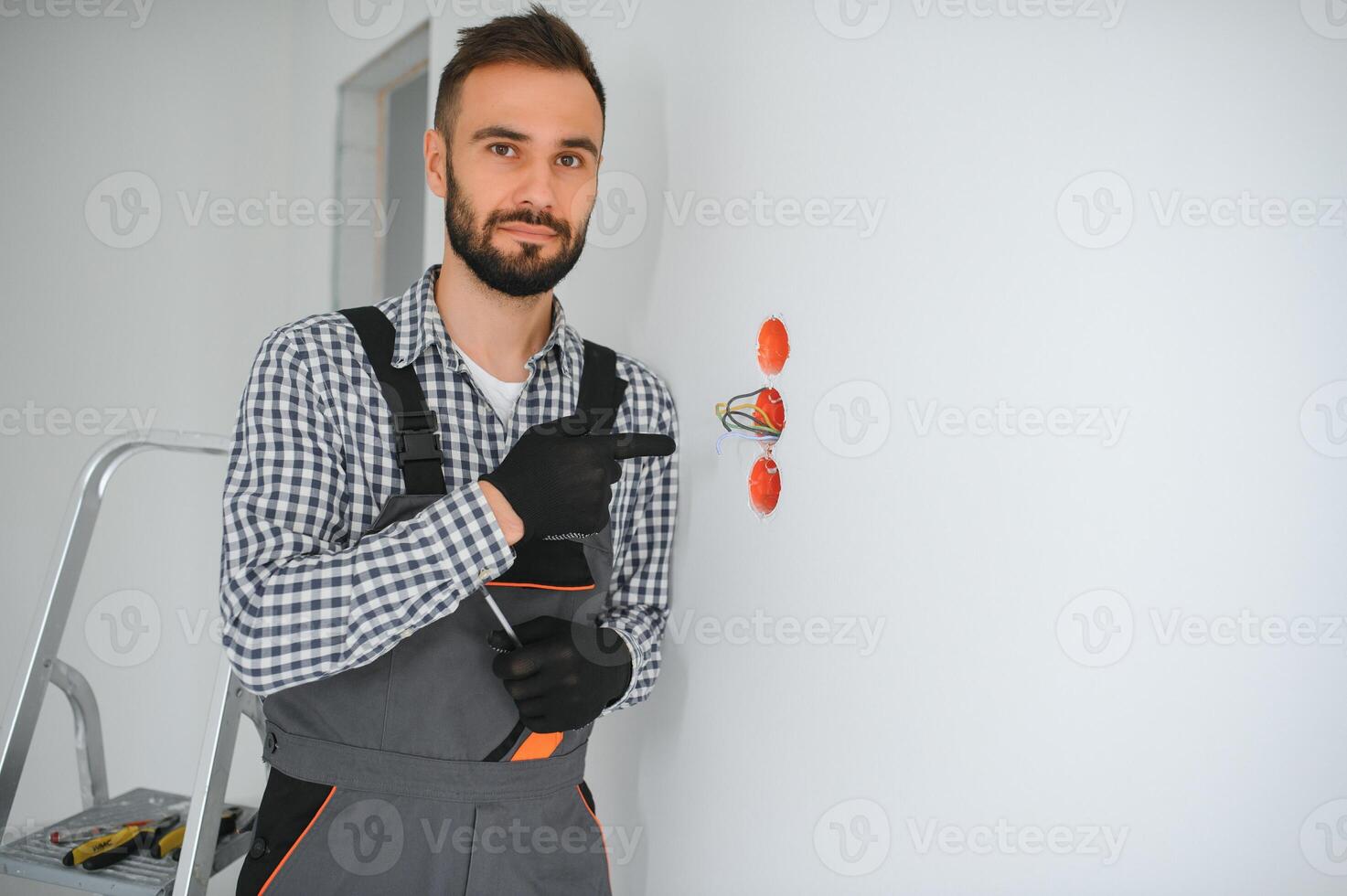 elettricista costruttore a opera, installazione di prese e interruttori. professionale nel tuta da lavoro con un elettricista attrezzo. contro il sfondo di il riparazione luogo foto