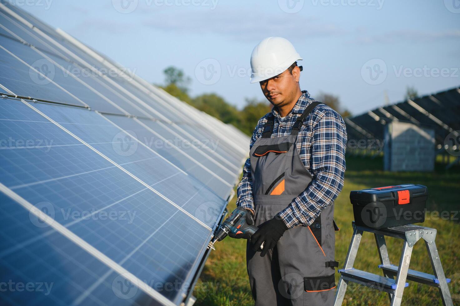 ritratto di giovane indiano uomo tecnico indossare bianca difficile cappello in piedi vicino solare pannelli contro blu cielo. industriale lavoratore solare sistema installazione foto