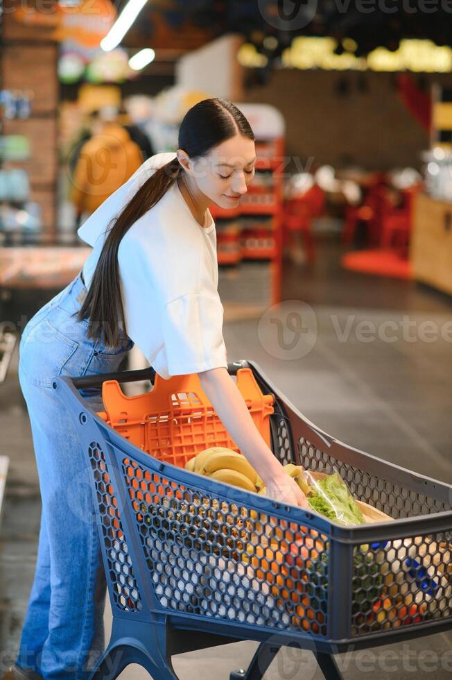 giovane sorridente contento donna 20s nel casuale Abiti shopping a supermercato memorizzare con drogheria carrello foto