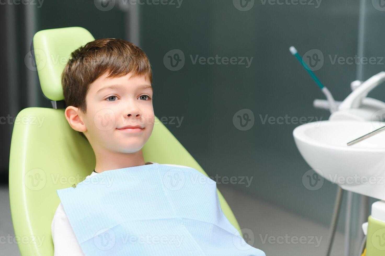 poco ragazzo si siede su dal dentista sedia nel bene umore dopo dentale procedure. giovane paziente con salutare denti foto