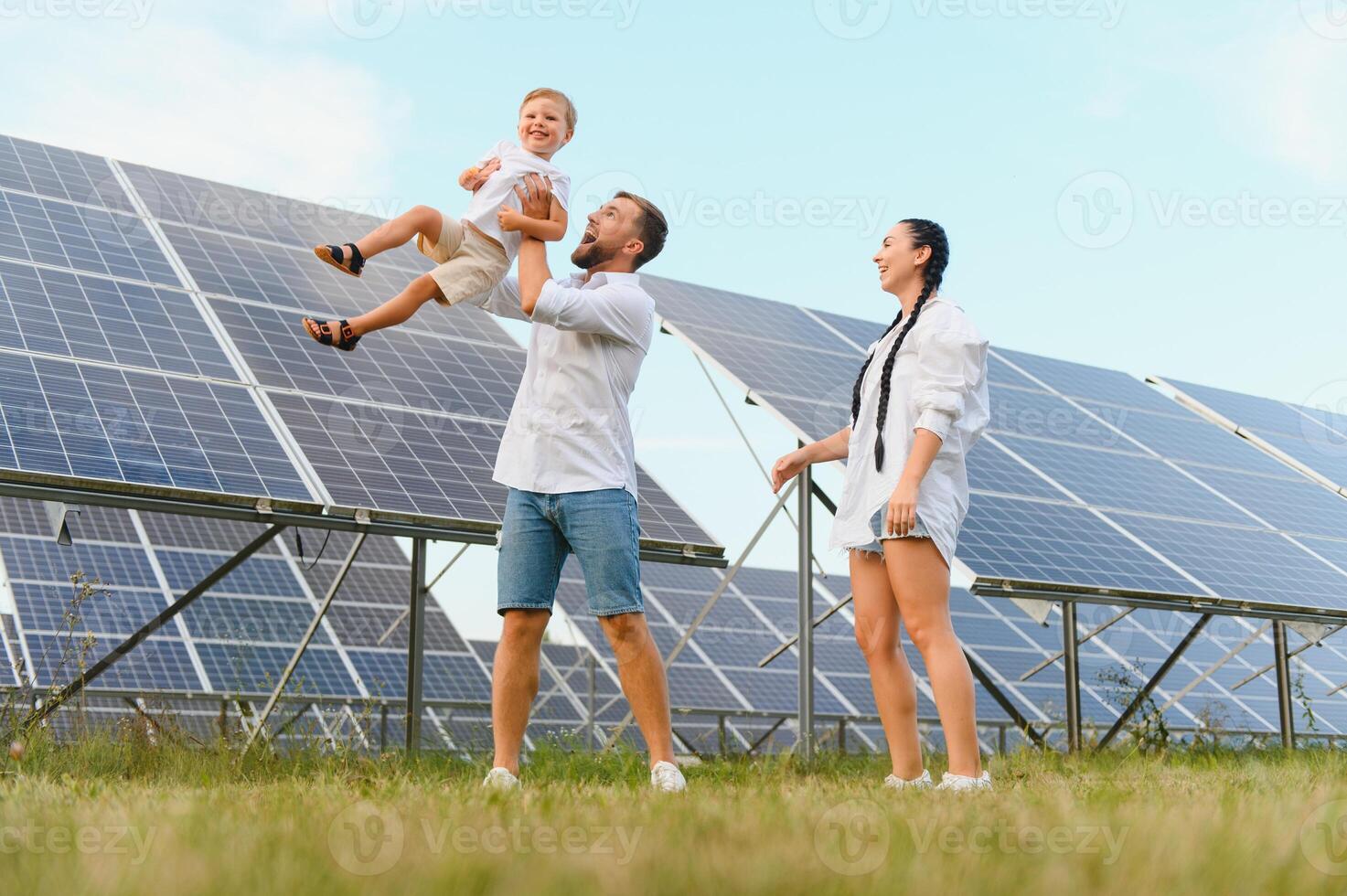 un' largo tiro di un' contento famiglia in piedi insieme e sorridente a telecamera con un' grande solare pannello nel sfondo foto