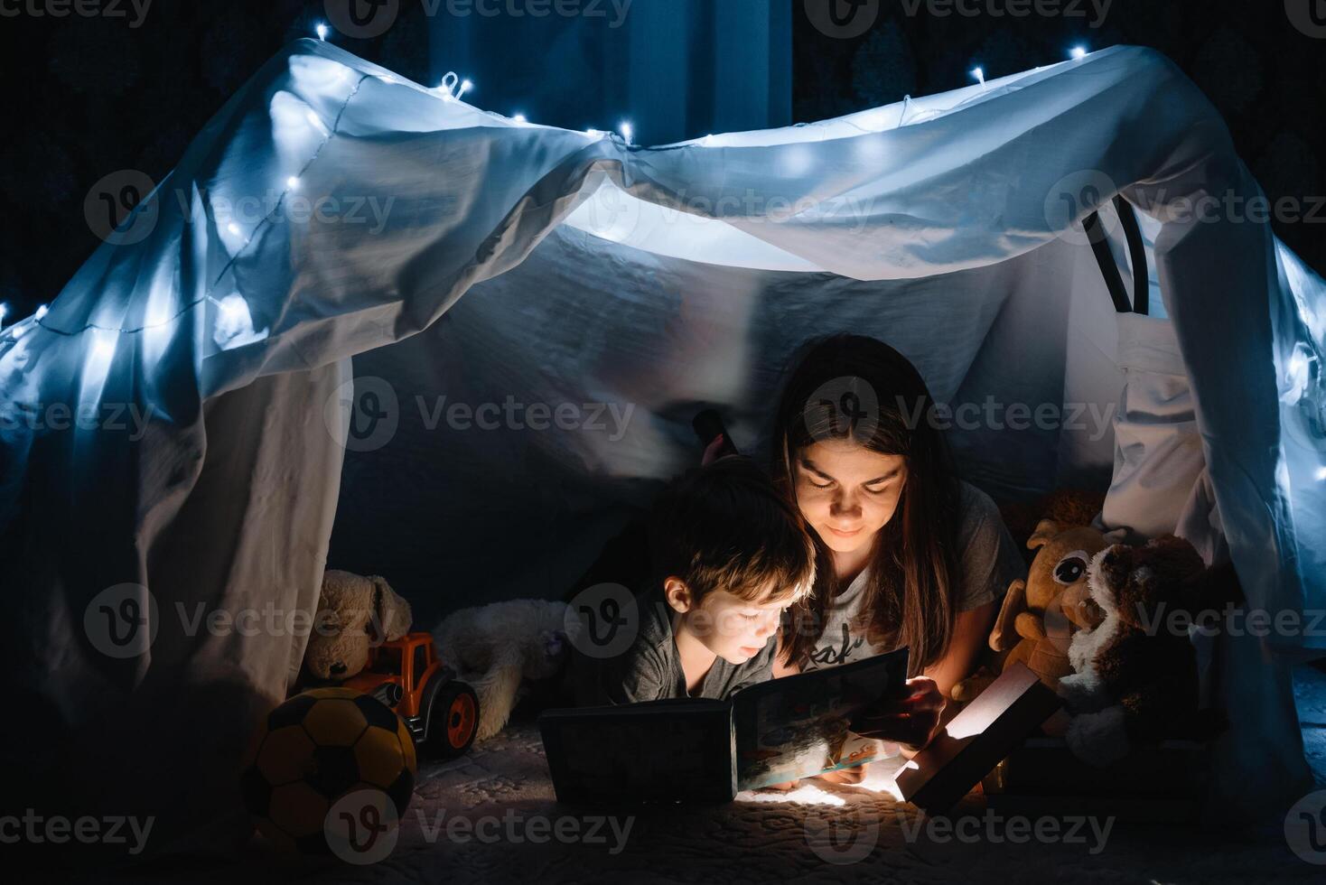 contento famiglia madre e bambino figlio lettura un' libro con un' torcia elettrica nel un' tenda a casa. famiglia concetto. foto