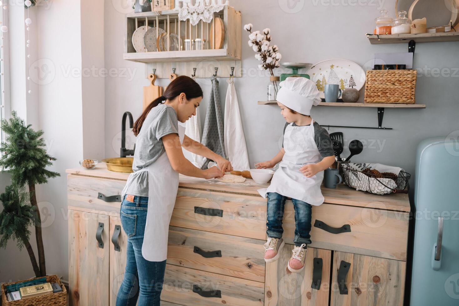 giovane contento mamma e sua bambino cucinare biscotti a casa nel il cucina. Natale fatti in casa Pan di zenzero. carino ragazzo con madre nel bianca uniforme e cappello cucinato cioccolato biscotti. foto