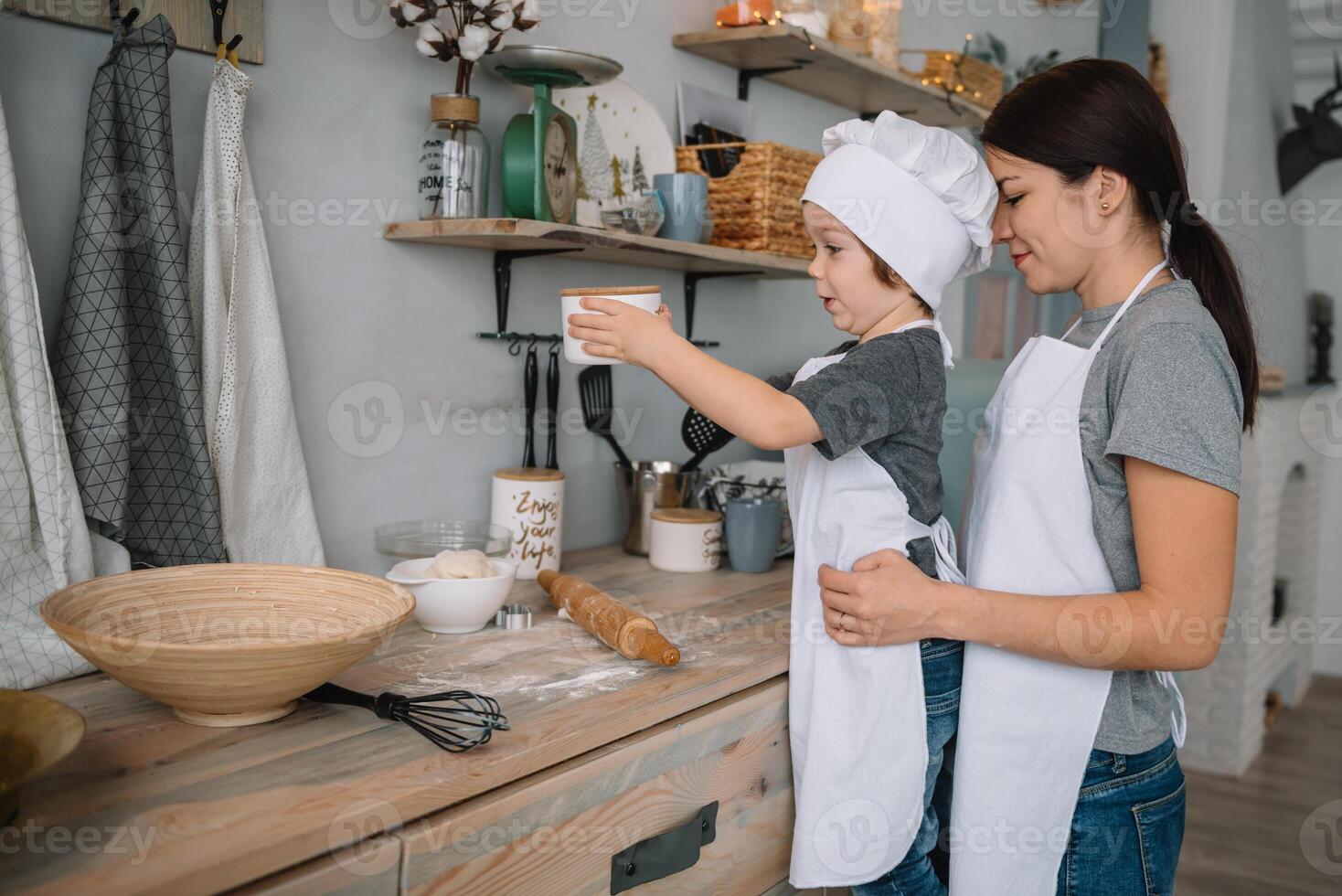 giovane contento mamma e sua bambino cucinare biscotti a casa nel il cucina. Natale fatti in casa Pan di zenzero. carino ragazzo con madre nel bianca uniforme e cappello cucinato cioccolato biscotti foto