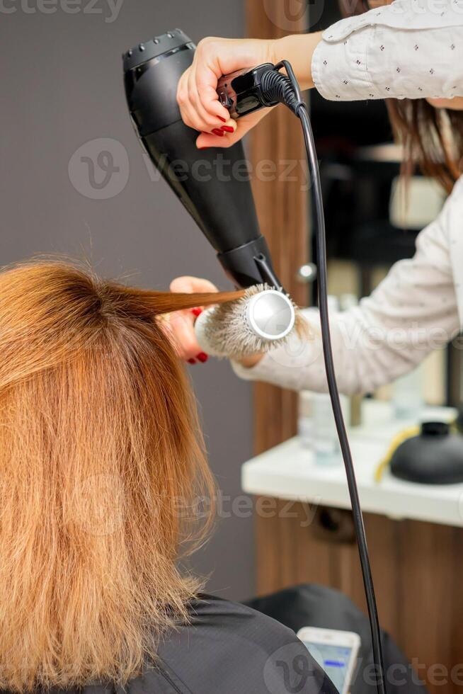 essiccazione capelli nel il capelli studio. femmina parrucchiere stilista si asciuga capelli con un' asciugacapelli e il giro spazzola rosso capelli di un' donna nel un' bellezza salone. foto