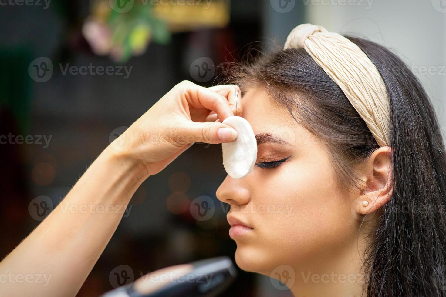 lato Visualizza di un' trucco artista coperture femmina occhio con un' cotone tampone utilizzando aerografo fabbricazione trucco fondazione su sua viso nel un' bellezza salone. foto