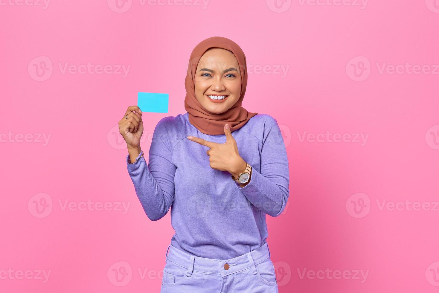 sorridente giovane donna asiatica che punta il dito sulla carta di credito su sfondo rosa foto