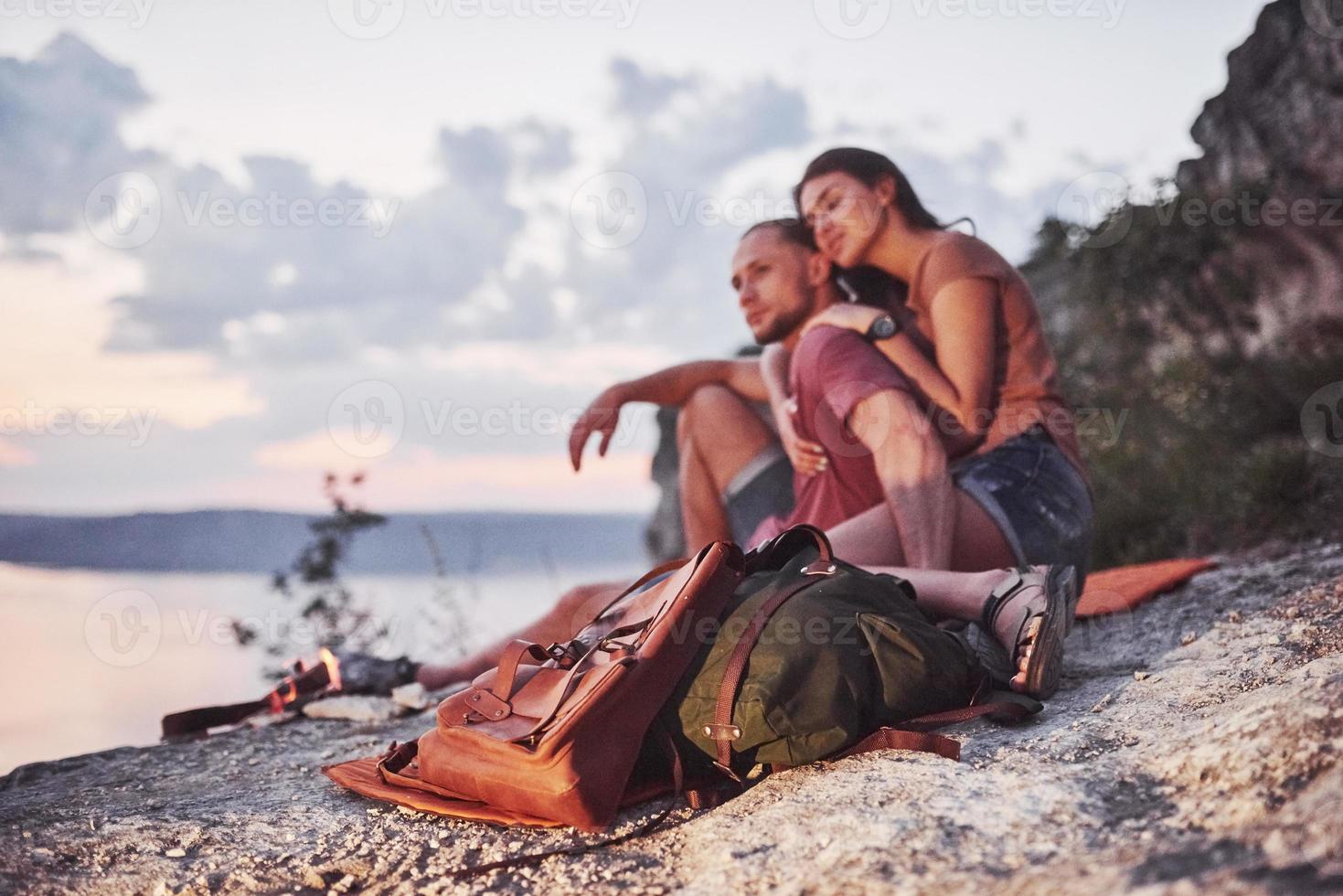 abbracciare la coppia con lo zaino seduto vicino al fuoco in cima alla montagna godendo della vista sulla costa di un fiume o di un lago. viaggiando lungo le montagne e la costa, la libertà e il concetto di stile di vita attivo foto