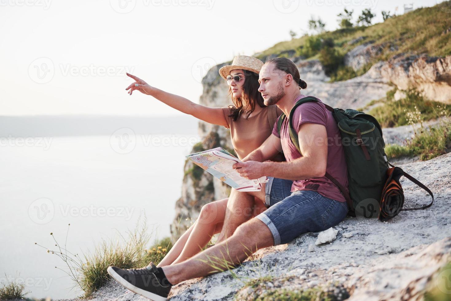 abbracciare la coppia con lo zaino seduto in cima alla montagna rocciosa godendo della vista sulla costa di un fiume o di un lago. viaggiando lungo le montagne e la costa, la libertà e il concetto di stile di vita attivo foto