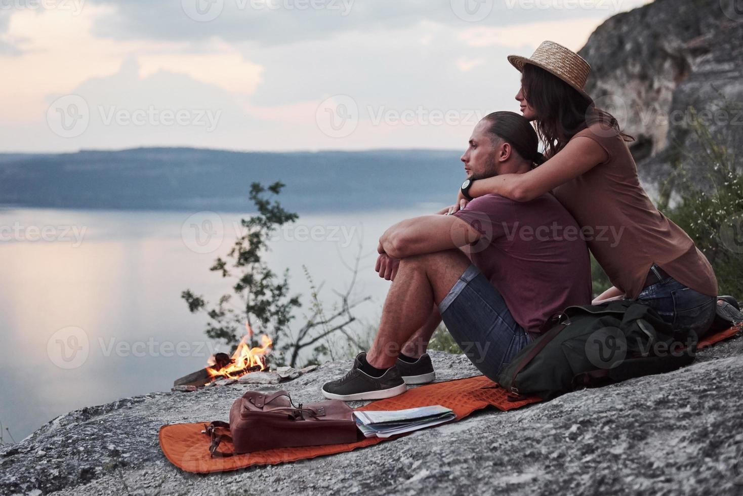 abbracciare la coppia con lo zaino seduto vicino al fuoco in cima alla montagna godendo della vista sulla costa di un fiume o di un lago. viaggiando lungo le montagne e la costa, la libertà e il concetto di stile di vita attivo foto