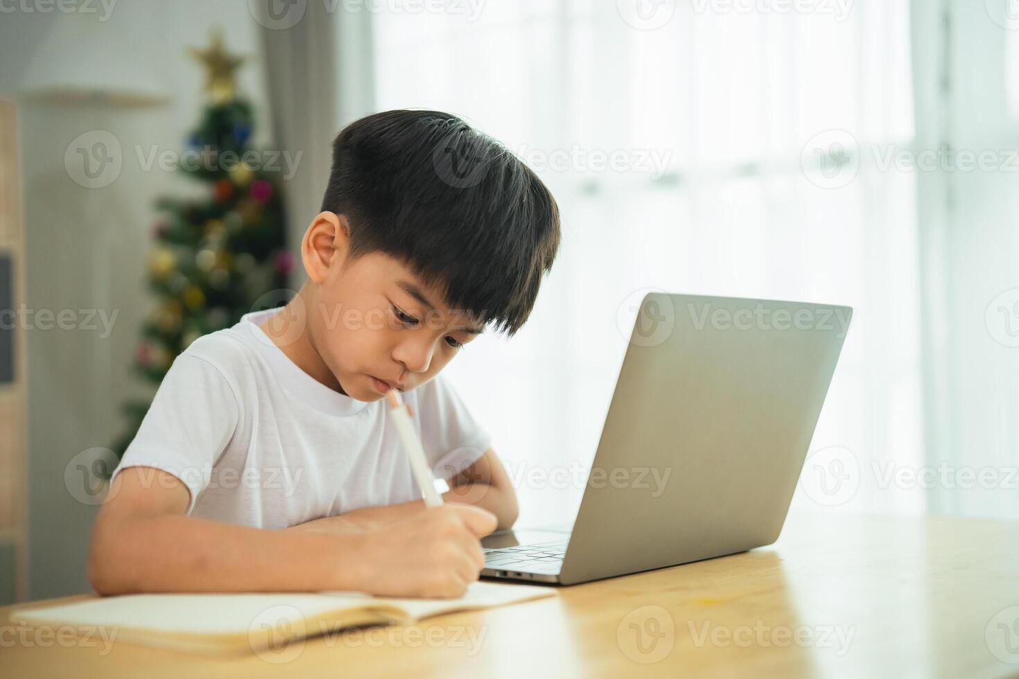 un' giovane ragazzo è seduta a un' tavolo con un' il computer portatile e un' matita. lui è scrittura qualcosa su un' pezzo di carta foto