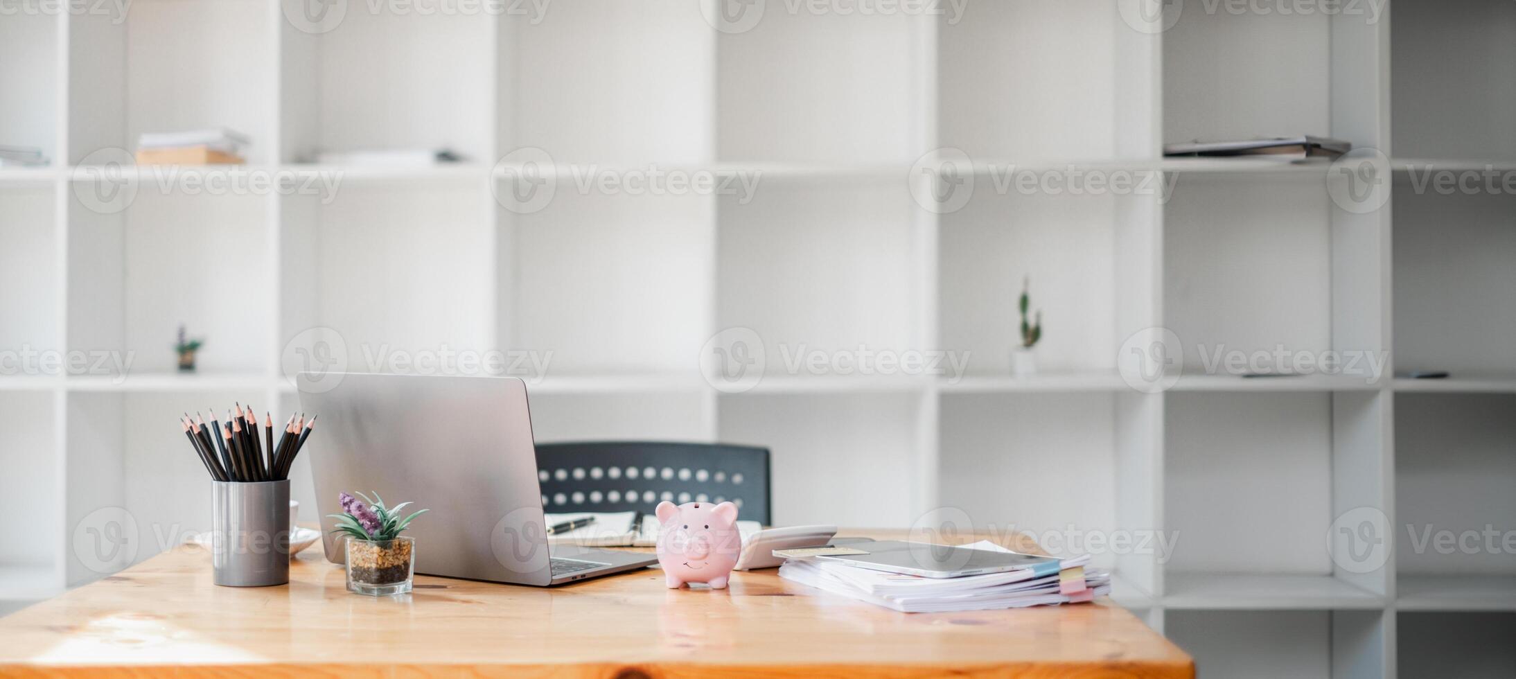 ordinato spazio di lavoro con un' computer portatile, colorato matite nel un' titolare, e un' porcellino banca, impostato contro un' fondale di bianca scaffali. foto