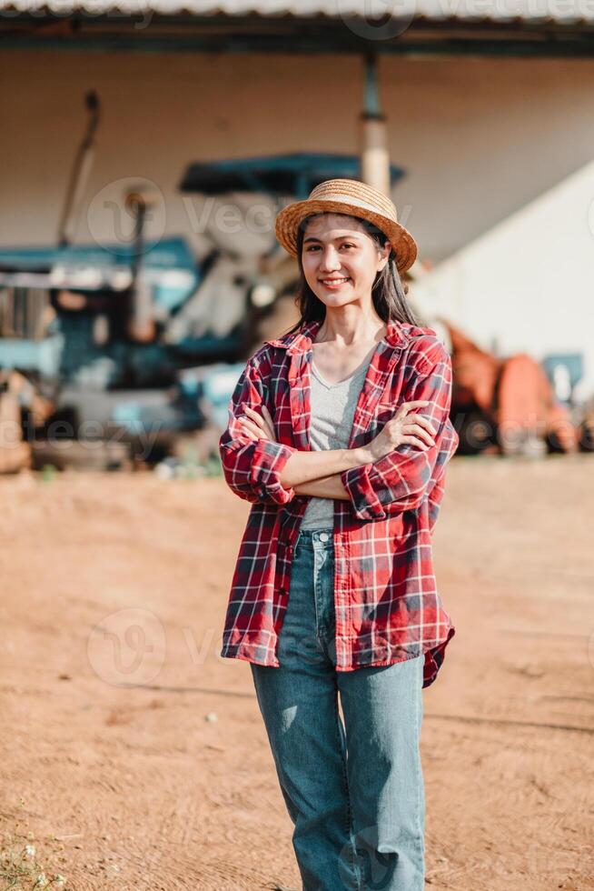 soddisfare contadino sta con braccia attraversato nel un' aia, indossare un' cannuccia cappello e plaid camicia, con agricolo macchinari nel il sfondo. foto