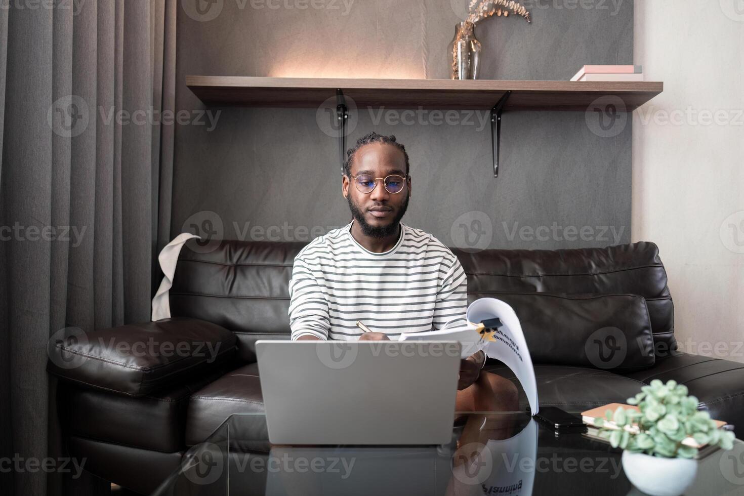 africano americano uomo Lavorando con il computer portatile computer a distanza mentre seduta a divano nel vivente camera. nero tipo fare libero professionista opera a casa ufficio foto