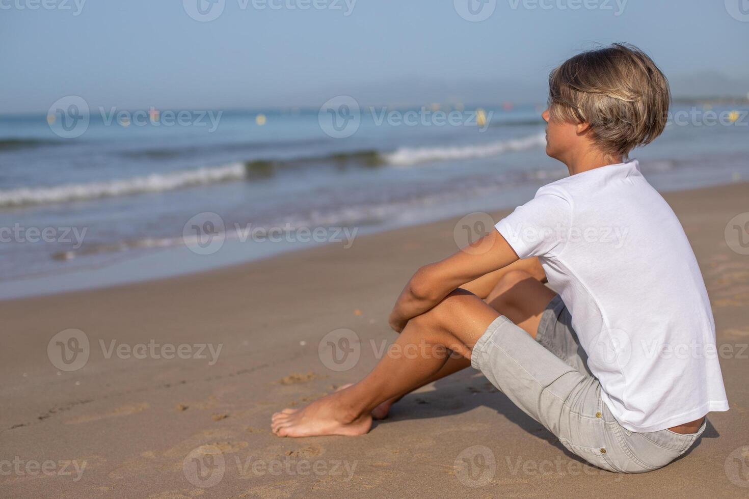 vicino su bello adolescente ragazzo nel bianca maglietta seduta a il spiaggia e godere un' estate vacanza guardare lontano.famiglia vacanze, vacanze, viaggio, viaggio concetto.lato Visualizza, copia spazio. foto