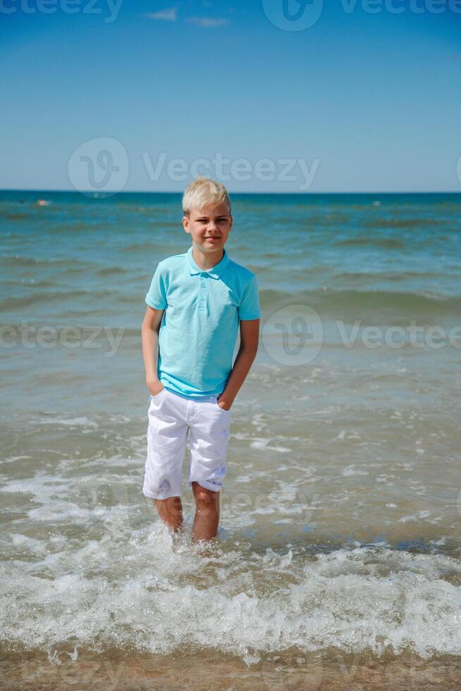 bello adolescente ragazzo nel bianca pantaloncini e un' blu maglietta sta nel il mare nel il acqua, e sembra per il telecamera. estate vacanza concetto.estate viaggio, famiglia vacanze, viaggio concetto.verticale foto. foto