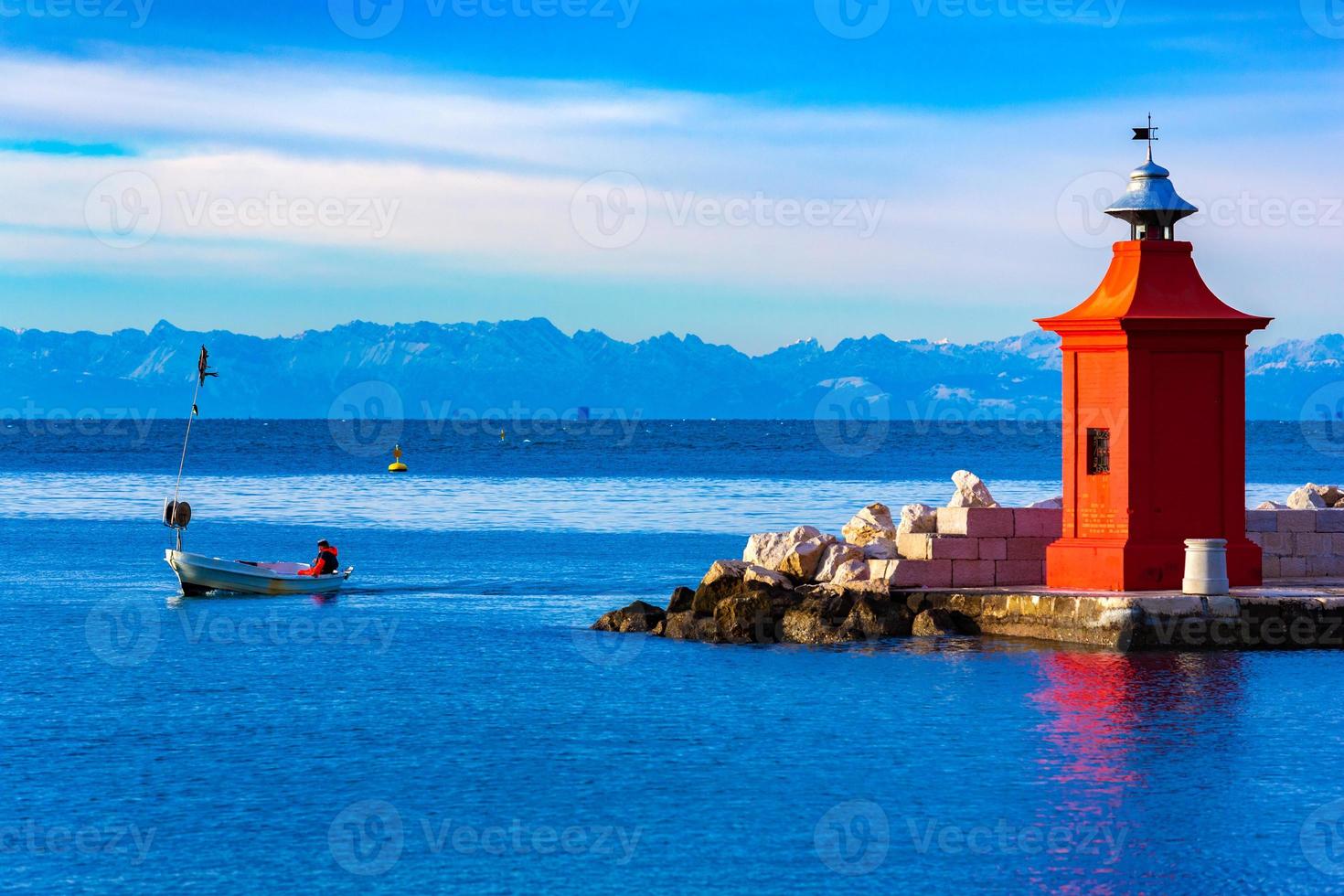 Vista del faro rosso su un molo, un pescatore su una barca e la bellissima catena montuosa all'orizzonte, pirano, slovenia foto