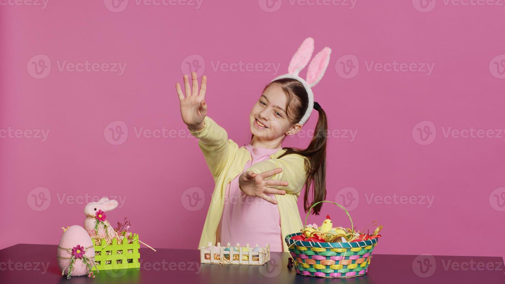 energico giovane ragazza con adorabile coniglietto orecchie agitando nel studio, detto Ciao e saluto qualcuno mentre lei crea Pasqua decorazioni. gioioso bambino piccolo in posa contro rosa sfondo. telecamera un. foto