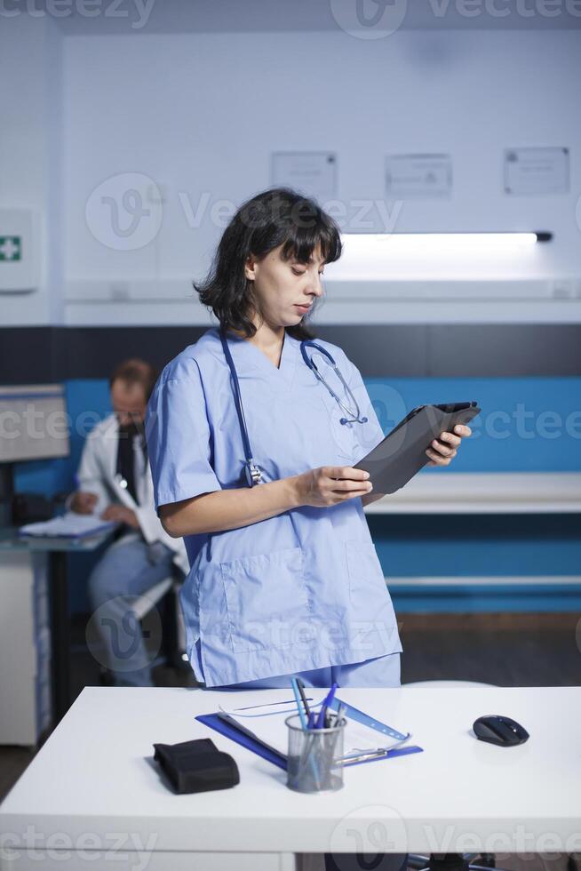 femmina medico lavoratore guardare a tavoletta nel un ufficio mentre in piedi Il prossimo per scrivania avendo penne, un' appunti, e un' topo. nel il sfondo si siede un' maschio medico Lavorando su assistenza sanitaria trattamenti. foto