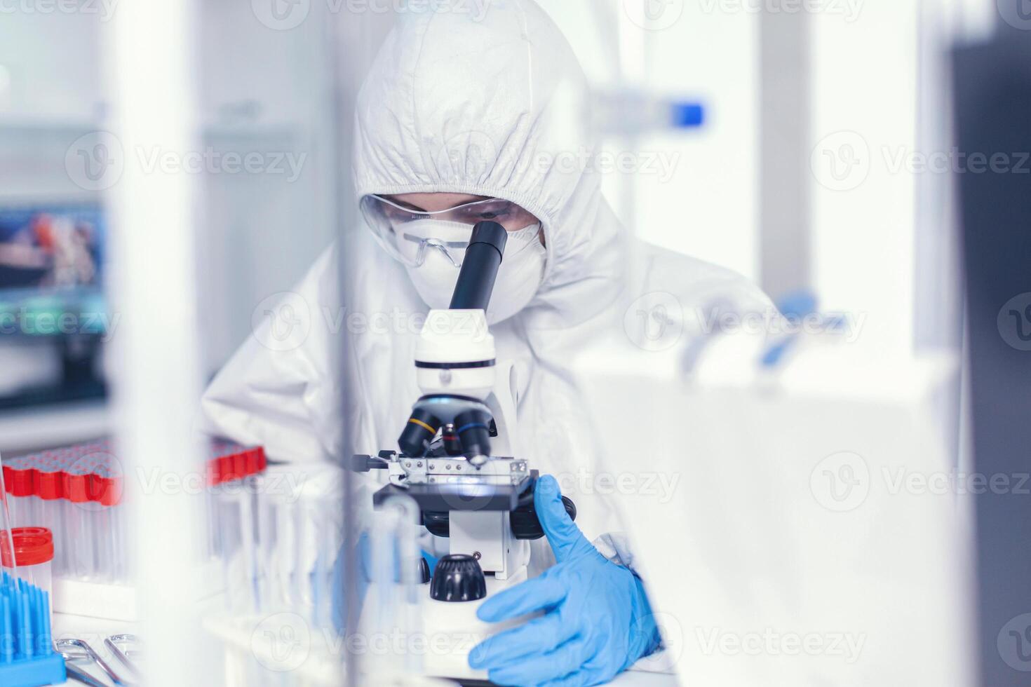 scienziato nel sterile protezione completo da uomo analizzando coronavirus guardare attraverso microscopio. chimico nel tuta l'esame vaccino Evoluzione utilizzando alto Tech ricercando diagnosi contro covid19 virus foto