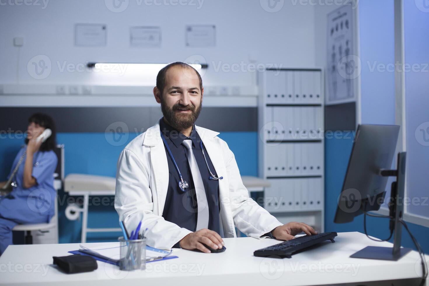 mezzo anziano medico sorridente a il suo scrivania nel un' medico ufficio. caucasico maschio medico appare per essere indossare un' bianca laboratorio cappotto, femmina infermiera è nel il sfondo su un' chiamata. ritratto sparo. foto