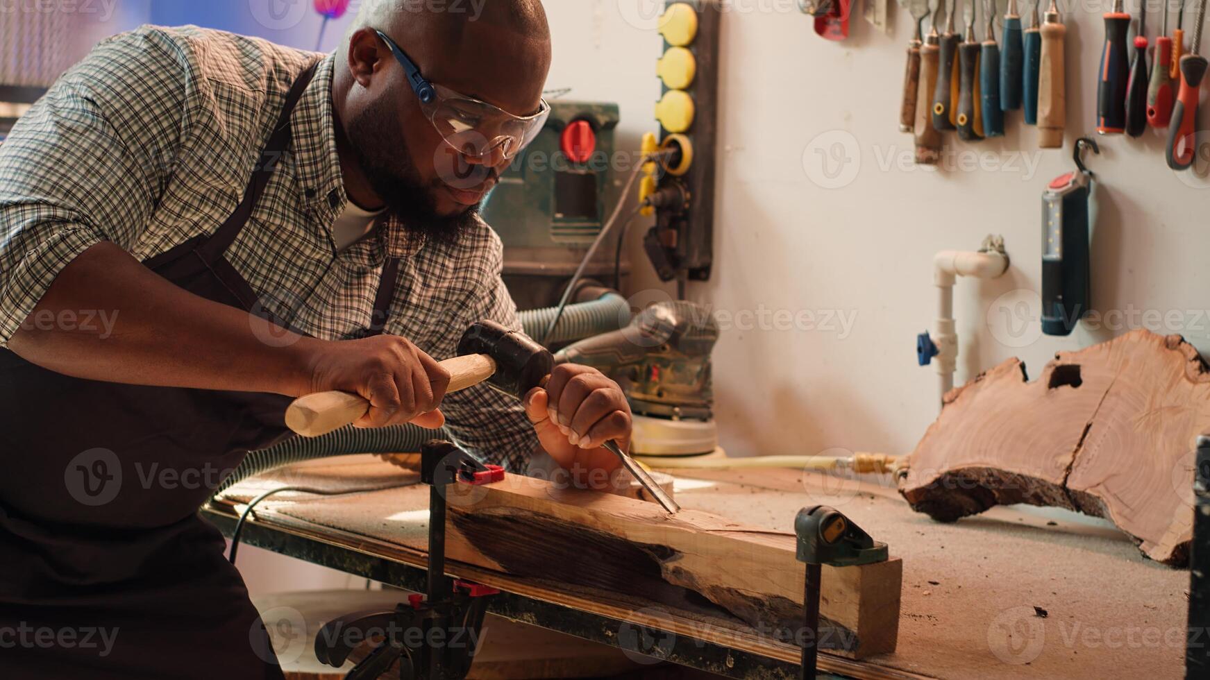 africano americano scultore modellare crudo legname utilizzando scalpello e martello nel carpenteria negozio, la creazione di legna arte, indossare sicurezza occhiali. artista fabbricazione legna sculture, incisione legname con Strumenti, telecamera un' foto