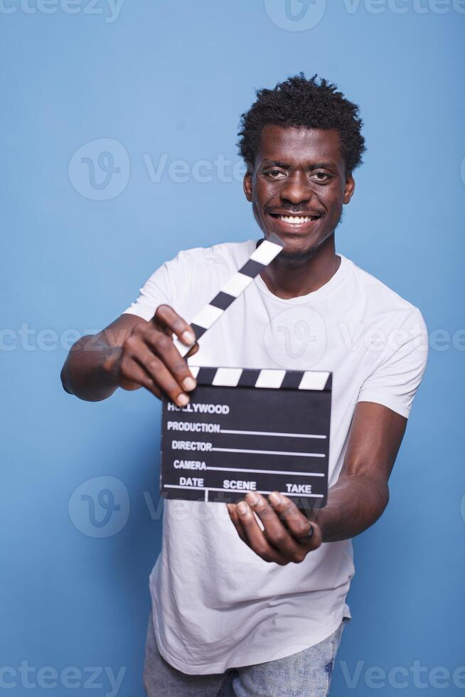 africano americano regista Tenere Aperto battacchio nel mani guardare fiducioso con un' Sorridi su il suo viso. nero film produttore con ciak in piedi orgoglioso nel davanti di isolato blu sfondo. foto