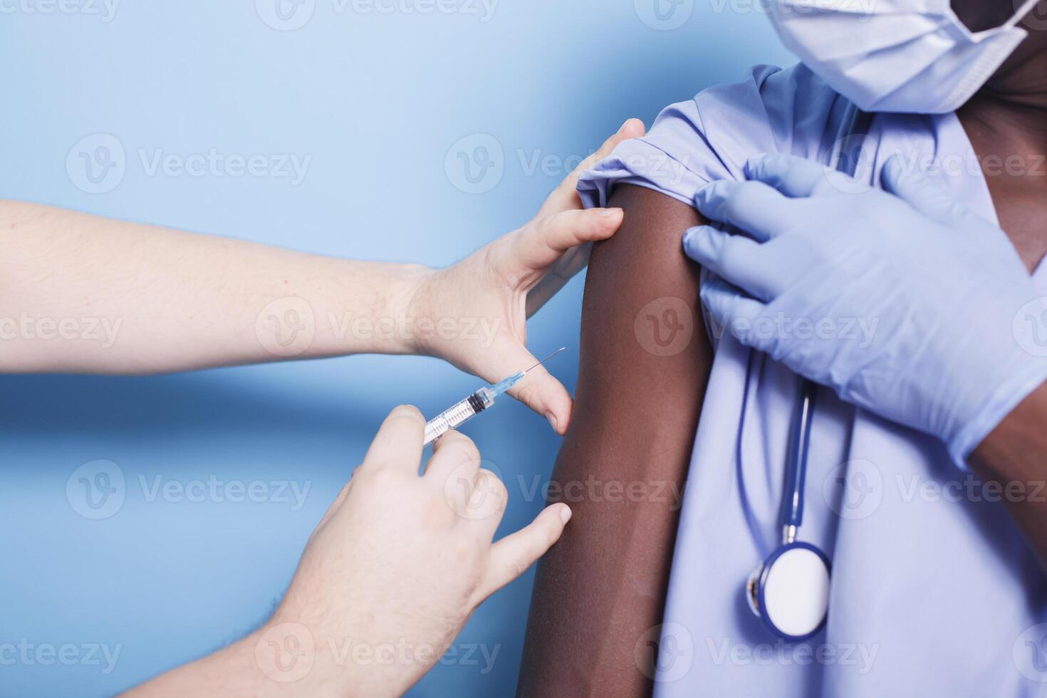 africano americano infermiera indossare viso maschera, ottenere vaccinati con siringa e ago. avvicinamento di medico assistente ricevente vaccino tiro a partire dal mani di medico per virus protezione. foto