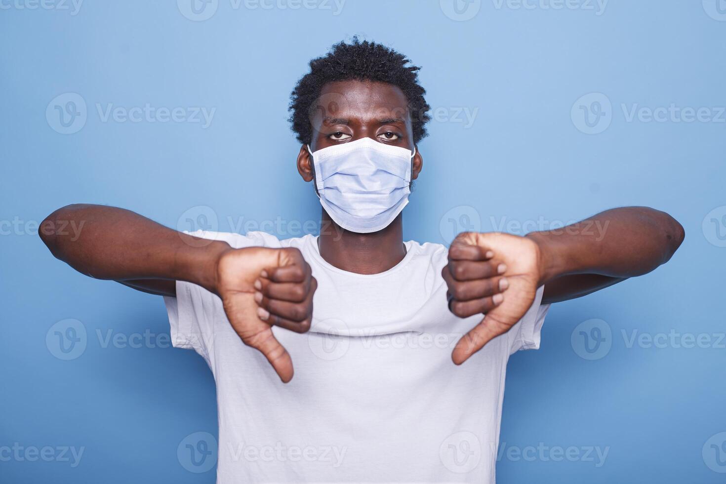 africano americano adulto mostrando pollici giù cartello con tutti e due mani. nero uomo Esprimere a gesti con il suo dita mentre guardare a telecamera e indossare viso maschera per proteggere a partire dal coronavirus epidemico. foto