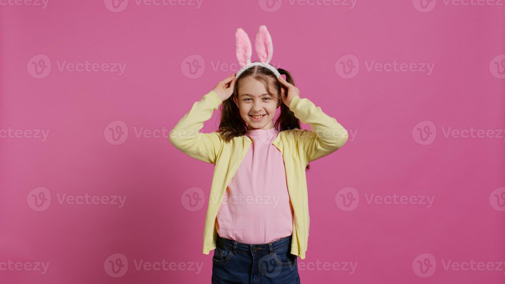adorabile carino bambino mettendo coniglietto orecchie e agitando a telecamera, godendo Pasqua Domenica celebrazione contro rosa sfondo. sorridente allegro scolara con trecce detto ciao. telecamera b. foto