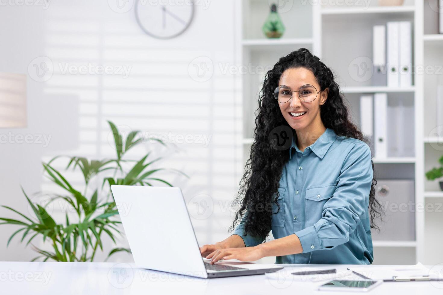 allegro ispanico donna nel un' casa ufficio ambientazione, sorridente a il telecamera mentre Lavorando su sua il computer portatile con impianti e un' orologio nel il sfondo. foto