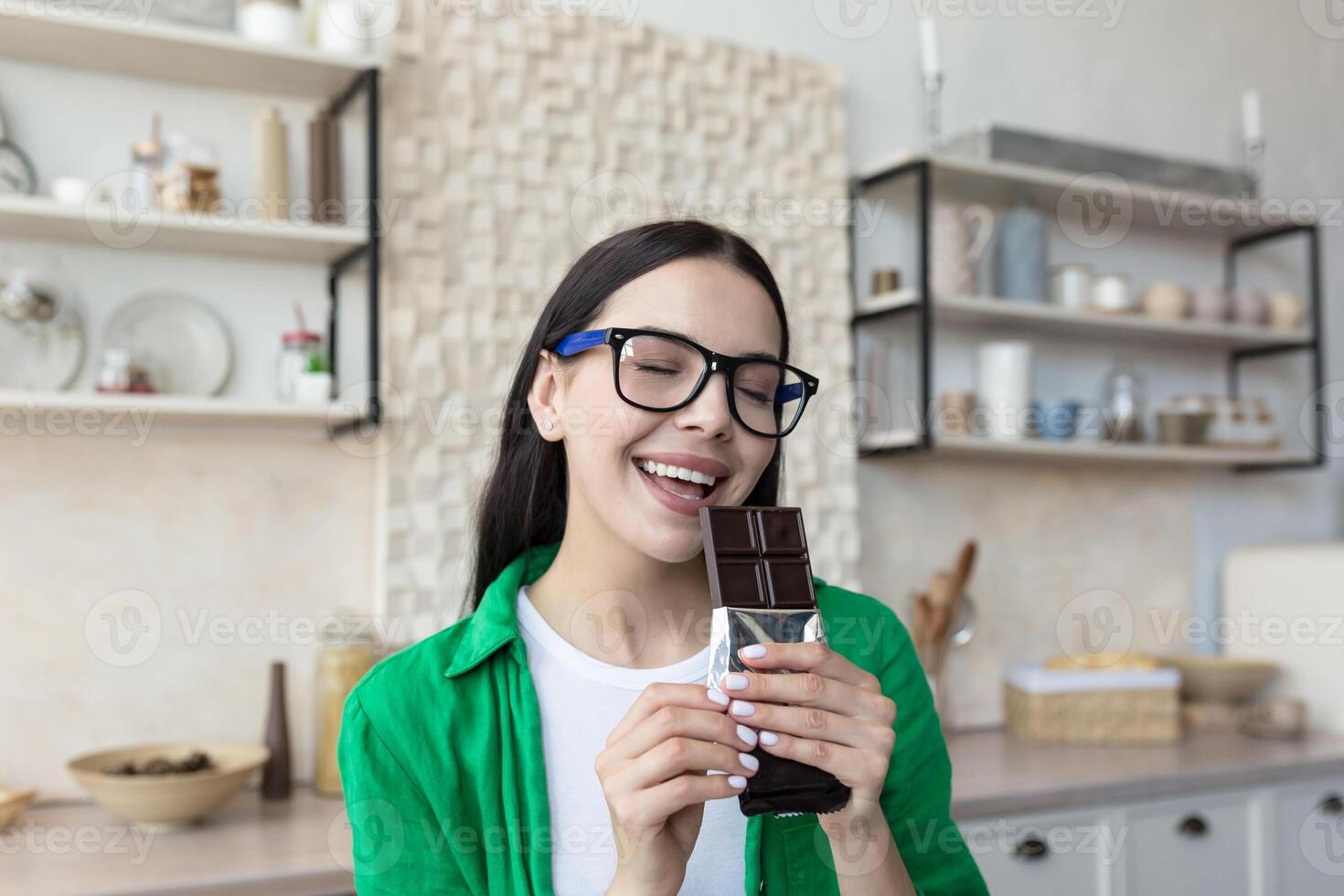 un' donna mangiare un' bar di buio cioccolato. in piedi a casa nel il cucina, occhi Chiuso, godendo foto