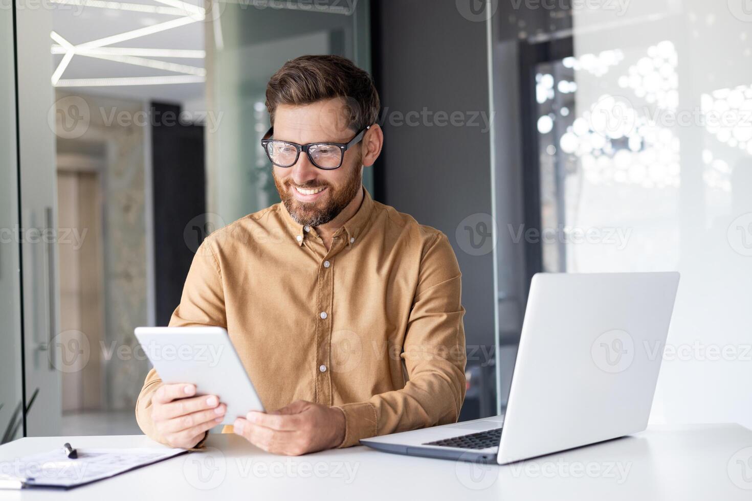un' giovane maschio programmatore sviluppatore lavori nel il ufficio a il tavolo a il il computer portatile e detiene un' tavoletta nel il suo mano. foto