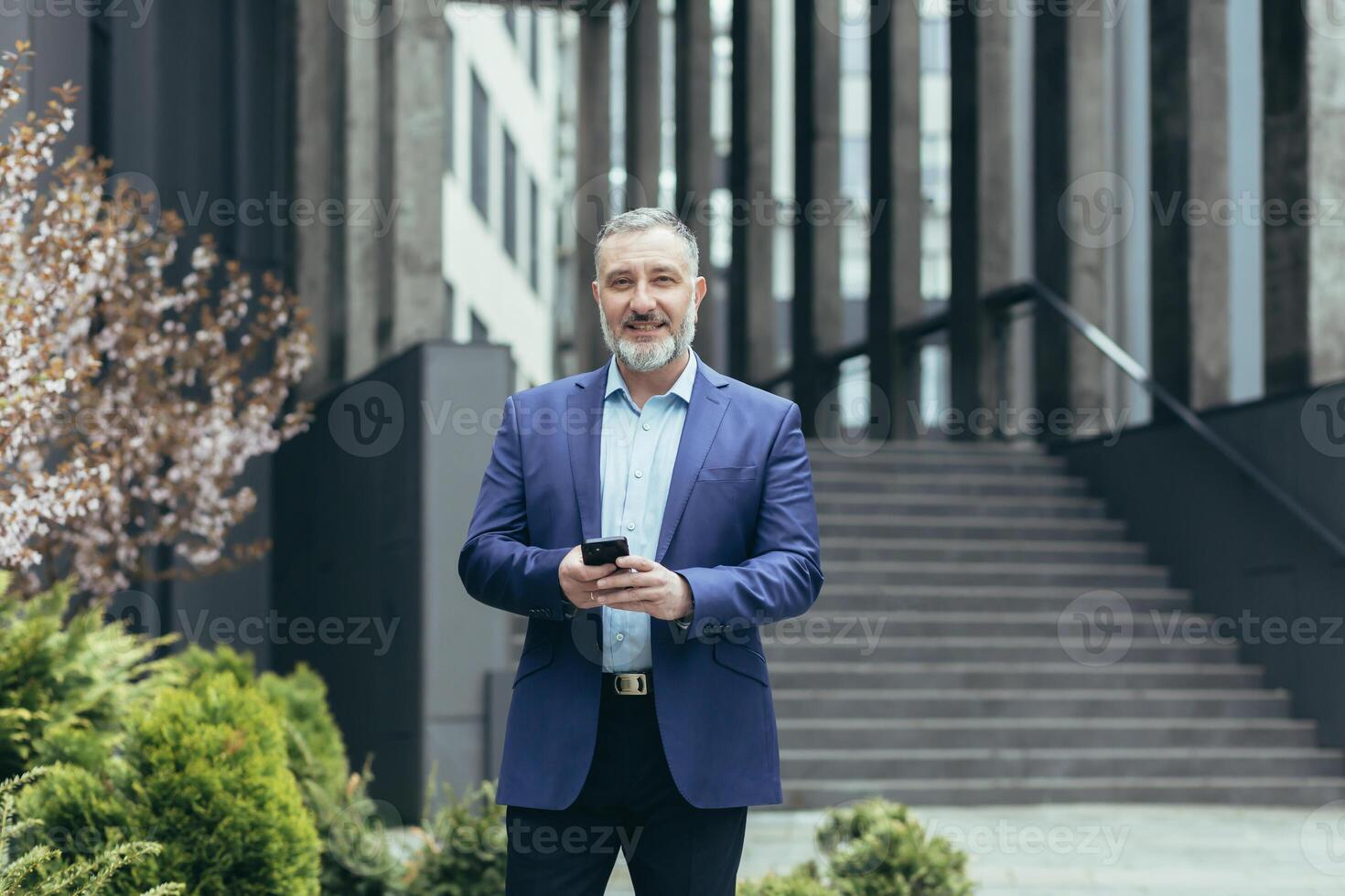 anziano bello dai capelli grigi uomo nel un' completo da uomo va per un' attività commerciale incontro, usi il Telefono su il strada, detiene esso nel il suo mani, quadranti. lui sembra a il telecamera, sorrisi. foto