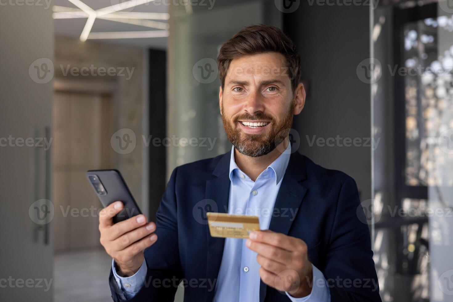 sorridente e riuscito giovane uomo uomo d'affari e banchiere in piedi nel ufficio, Tenere Telefono e credito carta, guardare con fiducia a telecamera. avvicinamento foto. foto