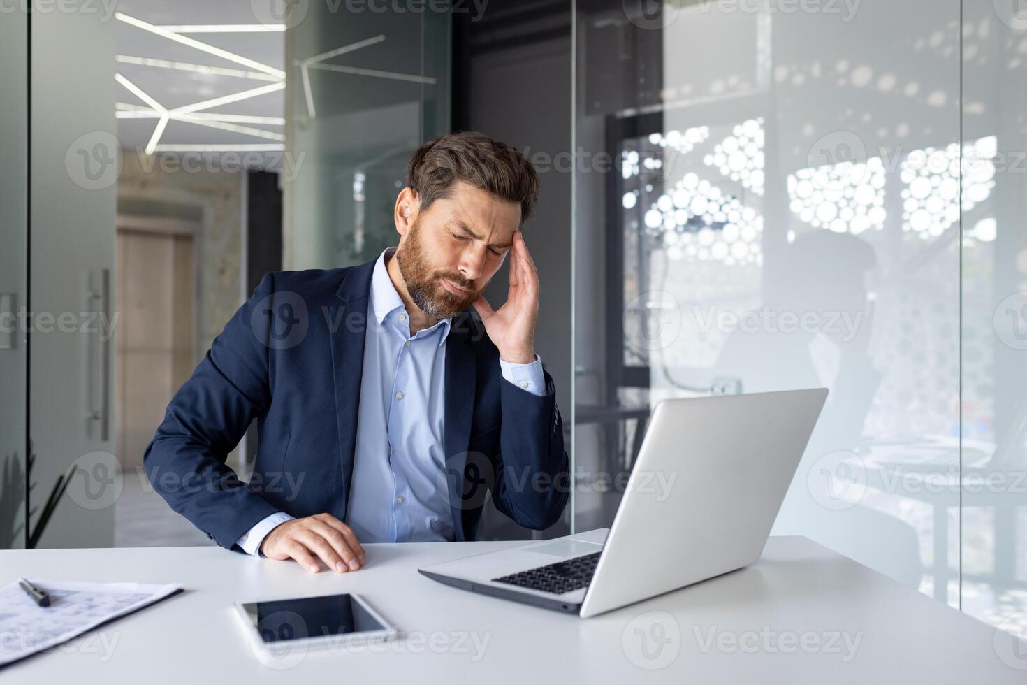 un' giovane uomo è seduta stanco e esausto a il posto di lavoro nel il ufficio e Tenere il suo testa con il suo mano, avendo un' acuto male alla testa, chiusura il suo occhi, riposo. foto