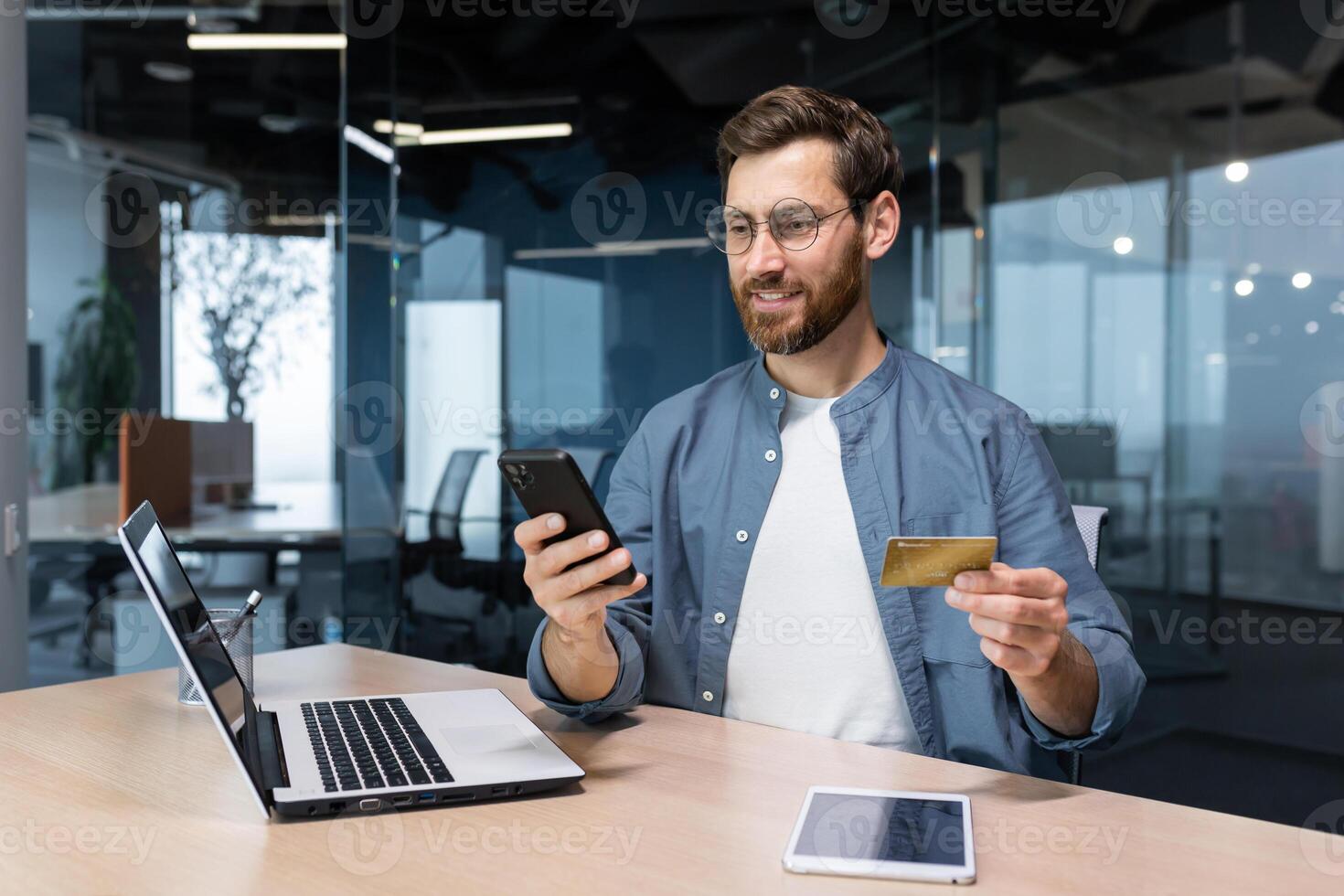 allegro e sorridente barbuto uomo d'affari fare in linea shopping e banca i soldi trasferimento, libero professionista Tenere banca credito carta e smartphone, uomo seduta a scrivania dentro ufficio. foto