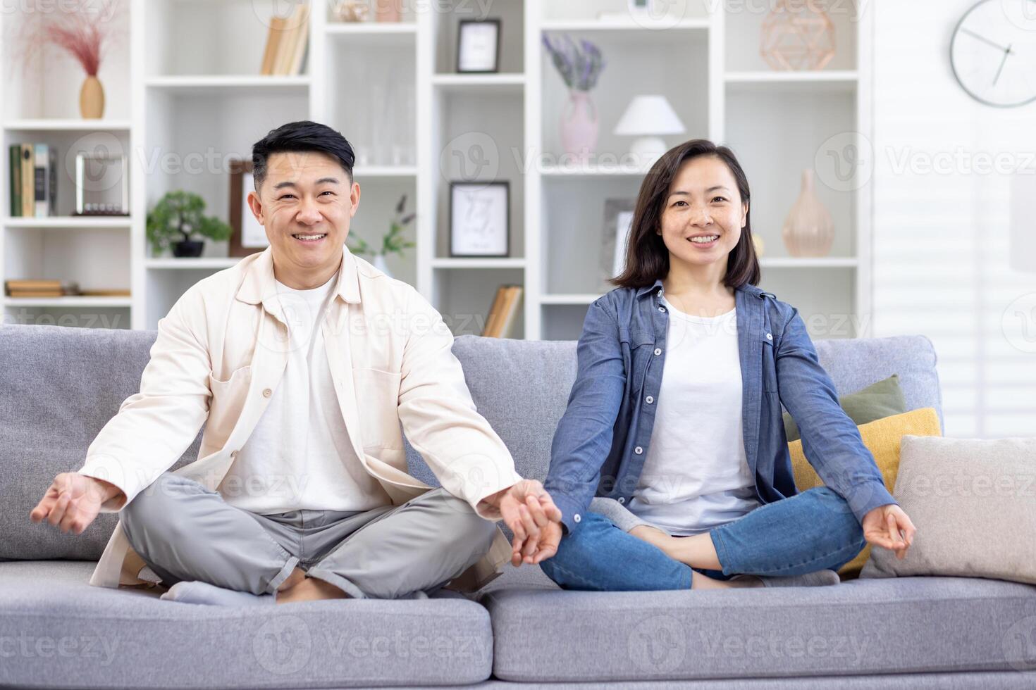 ritratto di un' giovane asiatico famiglia, un' coppia fare yoga a casa. un' uomo e un' donna siamo seduta su un' divano nel il loto posizione e sorridente a il telecamera. prendere cura di loro mentale Salute. foto