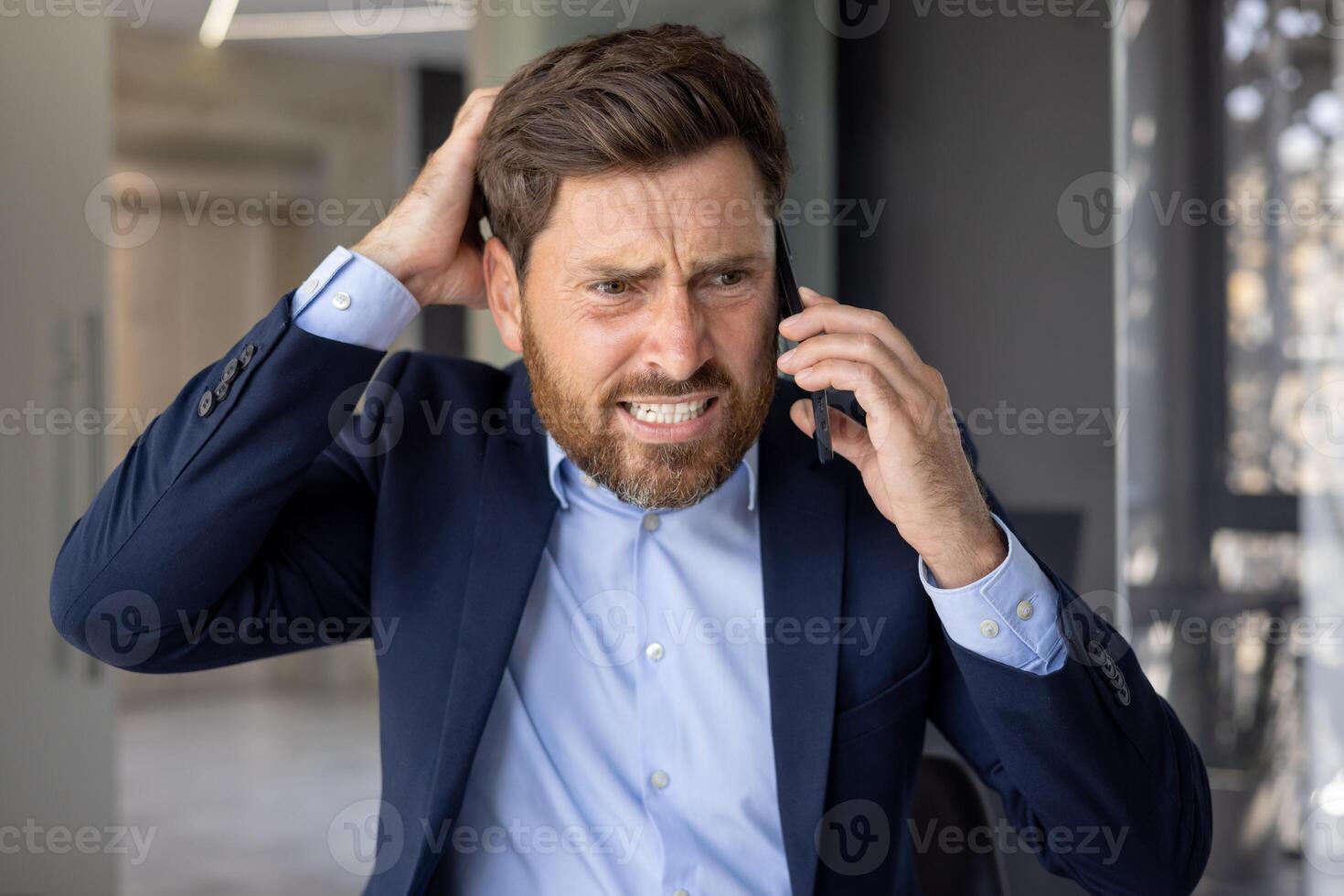 avvicinamento foto di un' giovane uomo d'affari uomo nel un' completo da uomo in piedi nel un ufficio centro, preoccupato parlando su il Telefono e Tenere il suo testa con il suo mano nel frustrazione, soluzione finanziario i problemi.