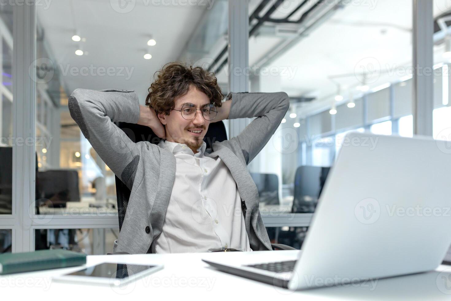 un' giovane fiducioso giovane uomo si siede nel il ufficio a il scrivania, sembra contento a il il computer portatile schermo, riposa con il suo mani dietro a il suo testa. foto