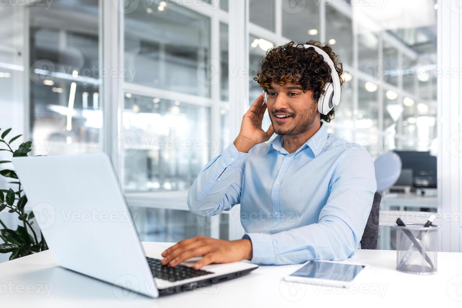 giovane riuscito programmatore dentro moderno ufficio Lavorando con computer portatile, uomo nel blu camicia sorridente e felice, ascoltando in linea musica nel cuffia, Audio libri e podcast, uomo d'affari soddisfatto opera. foto