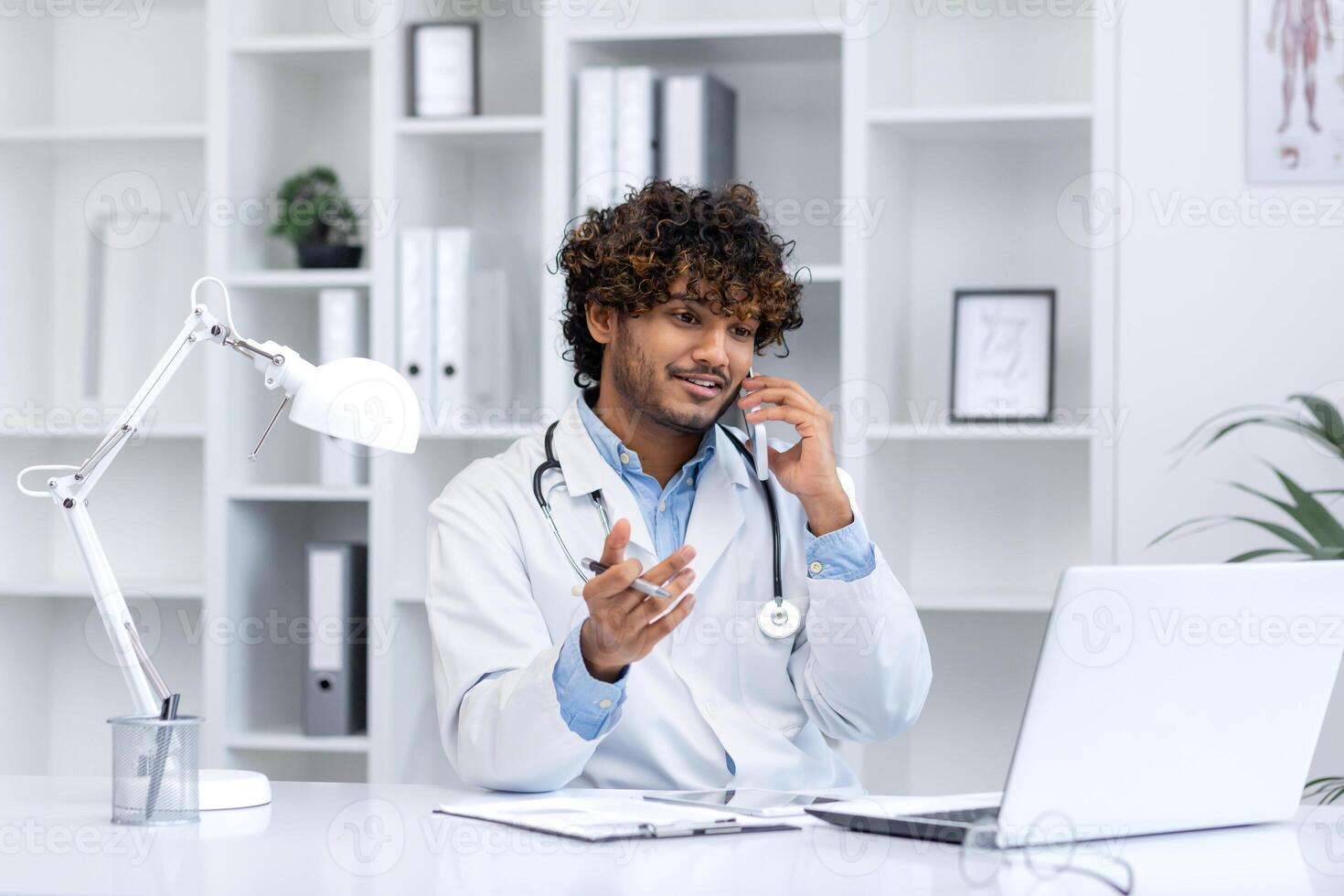 giovane riuscito ispanico medico dentro il clinica nel il medico ufficio con gioia comunica parlando su il Telefono, il clinica lavoratore si siede a il scrivania Lavorando con un' computer portatile, consulta pazienti. foto