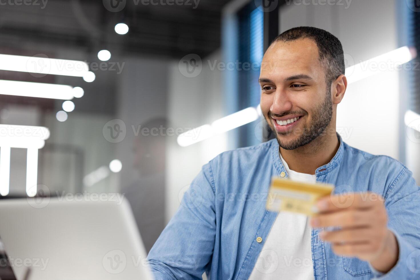 un' giovane adulto maschio Tenere un' credito carta, intenzione per rendere un in linea Acquista mentre Lavorando su il suo il computer portatile nel un' contemporaneo ufficio ambientazione. foto