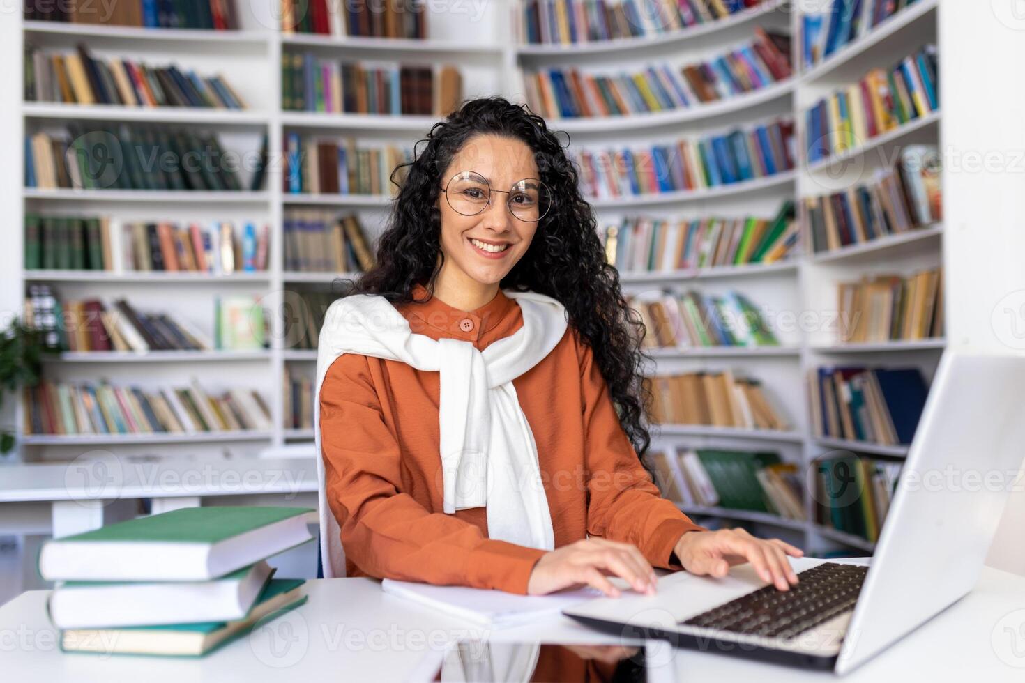 ritratto di allegro sorridente ragazza alunno, donna guardare a telecamera seduta studiando con computer portatile, ispanico donna indossare bicchieri studiando in linea nel Università biblioteca tra bicchieri utilizzando il computer portatile. foto