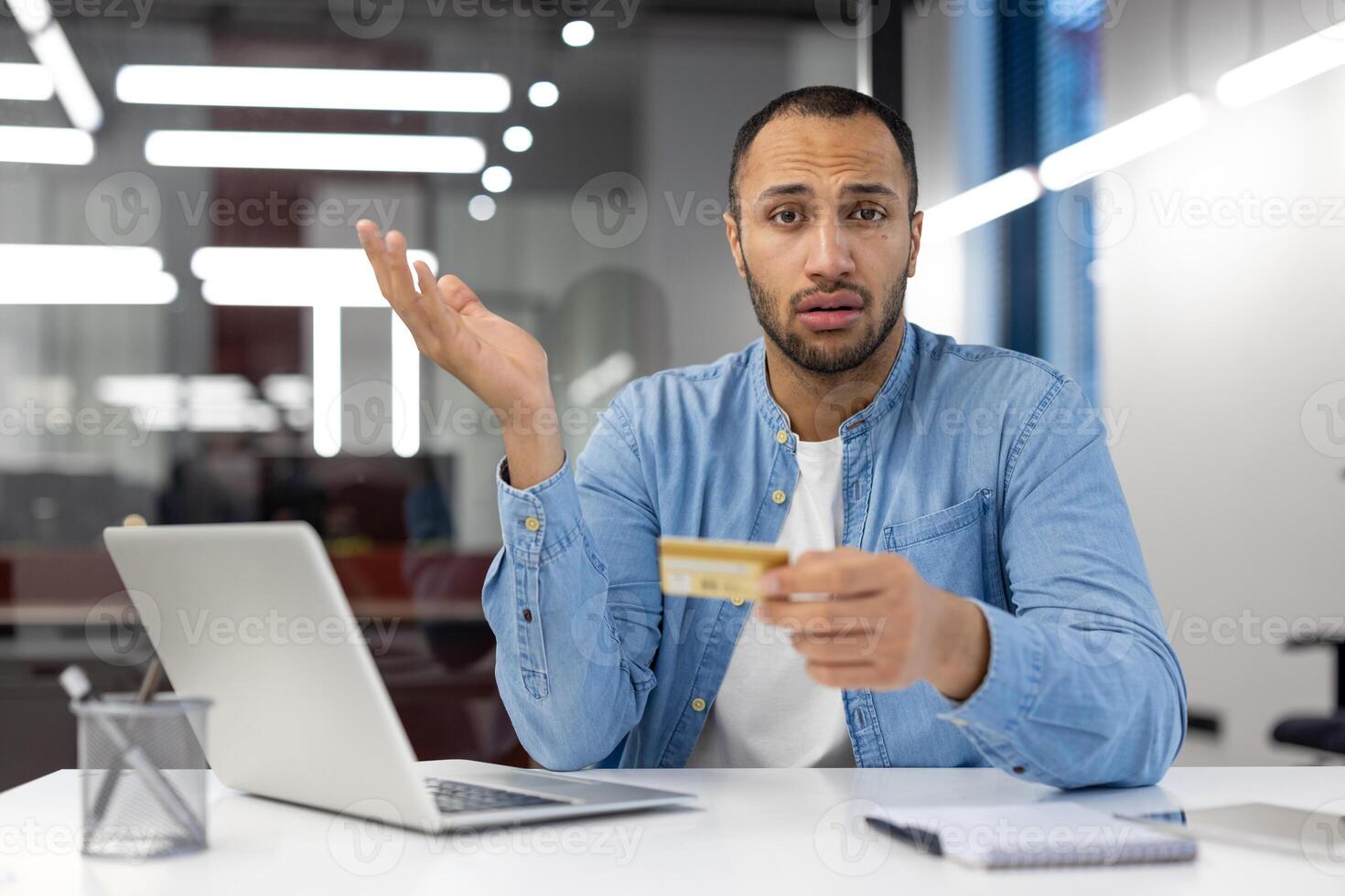 un' uomo è seduta a un' scrivania con un' il computer portatile e un' credito carta nel il suo mano. lui sembra confuso e è Tenere il carta su per il suo viso foto
