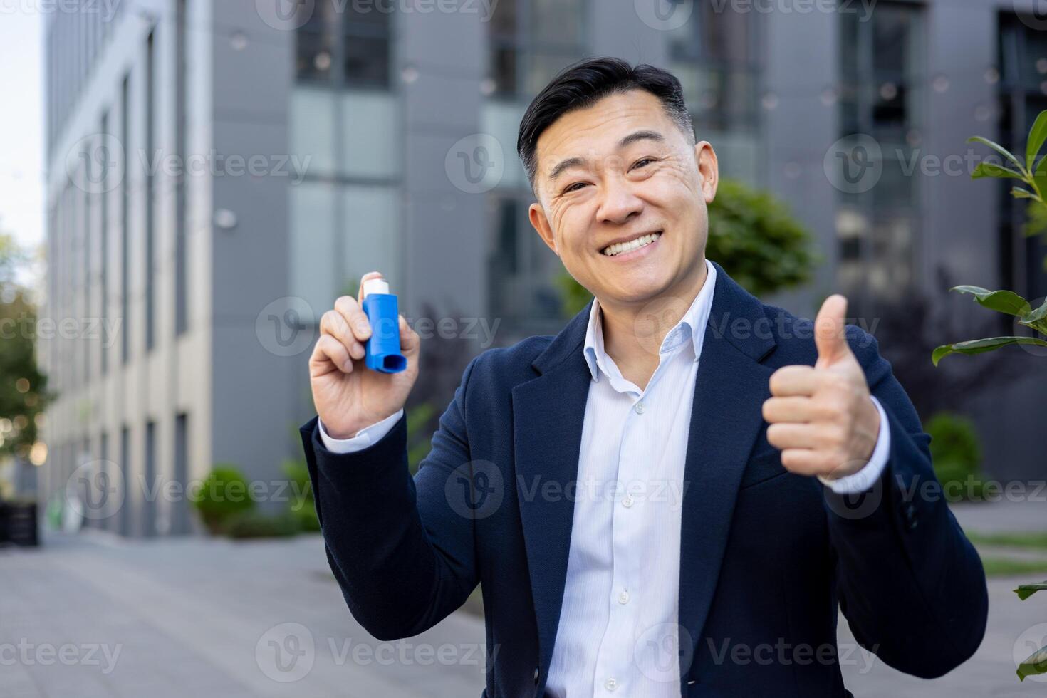 ritratto di un' sorridente giovane asiatico uomo nel un' attività commerciale completo da uomo seduta su un' panchina vicino un ufficio costruzione, Tenere un inalatore nel il suo mano e mostrando il super cartello con il suo dito per il telecamera. foto